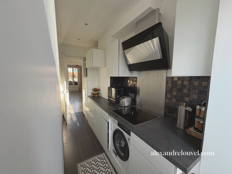 Petite cuisine moderne avec appareils électroménagers, comptoir noir et mur de carreaux décoratifs, vue vers une fenêtre lumineuse.