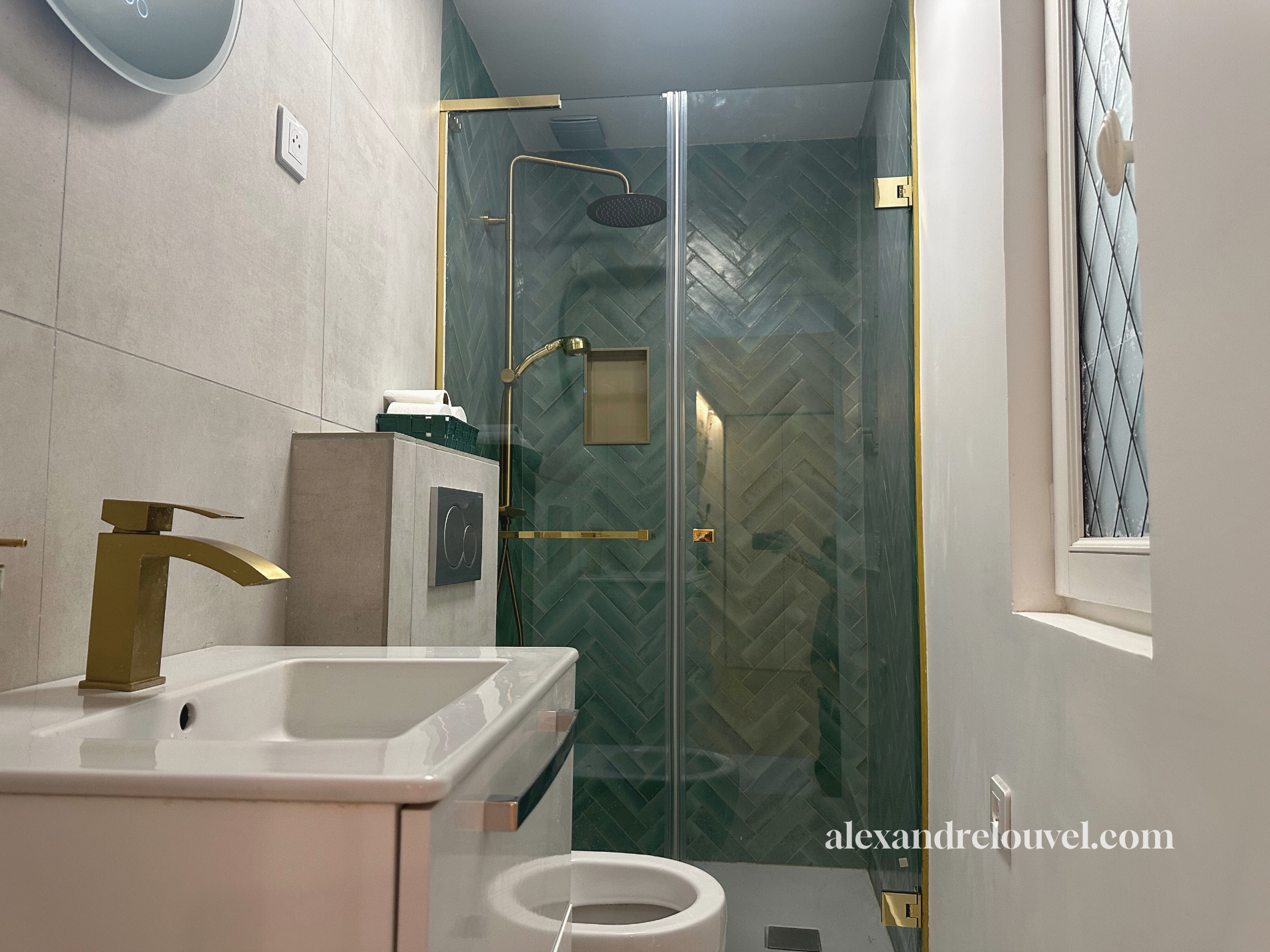 Salle de bain moderne avec douche en verre, mur en carrelage vert en motif en chevrons, évier blanc avec robinet en or, fenêtre avec grille, et un sol en carrelage beige.