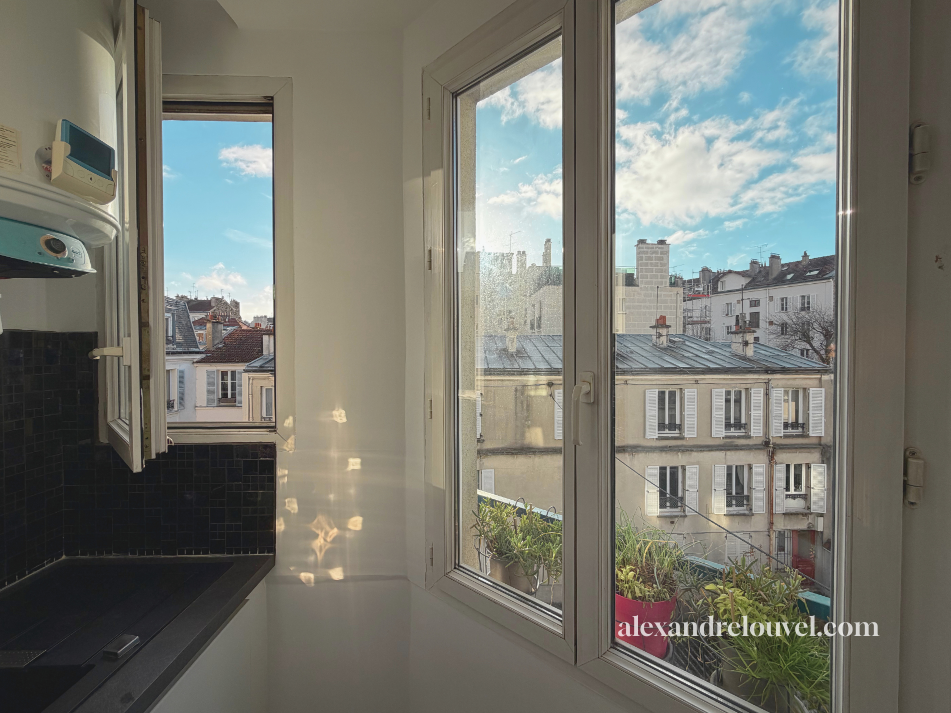 Vue depuis une fenêtre d'une cuisine urbaine avec des bâtiments résidentiels, une fenêtre ouverte, des plantes en pot sur un balcon, ciel en partie nuageux, site alexandrelouvel.com.