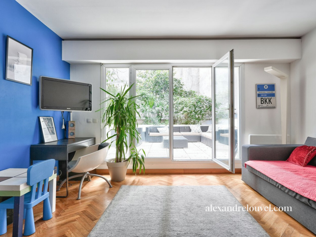 Salon moderne avec un mur bleu, un canapé gris avec un coussin rouge, une grande plante verte, un téléviseur mural, un bureau noir, un tableau et un balcon avec meubles extérieurs et plantes.