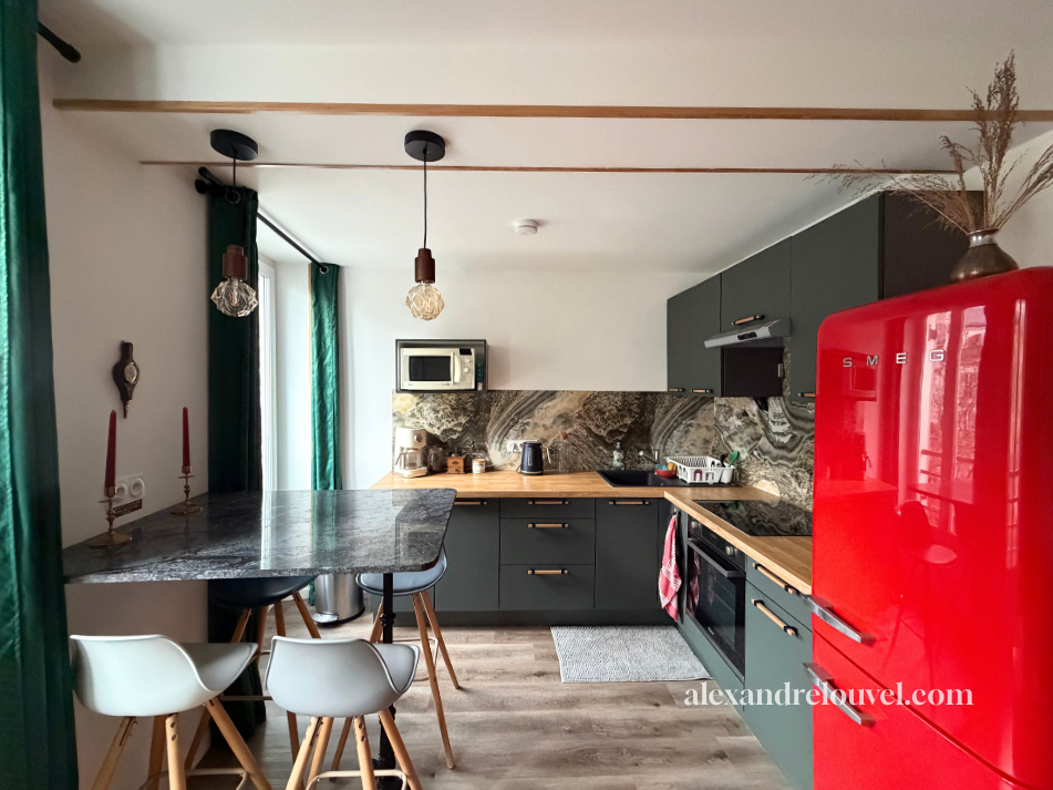Cuisine moderne avec armoires gris foncé, comptoir en bois et plan de travail en marbre, réfrigérateur rouge SMEG, gadgets de cuisine, luminaire suspendu au plafond, fenêtre avec rideaux verts, table en marbre avec trois chaises, décor avec candélabres, micro-ondes, appareils électroménagers et vase avec des plantes.