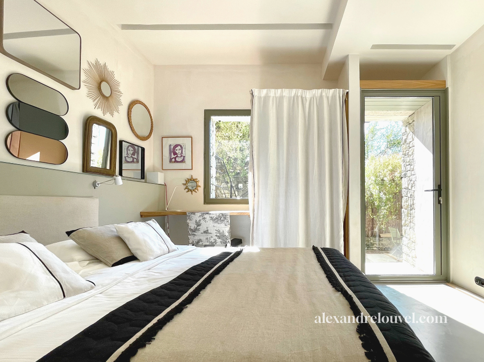 Chambre lumineuse avec un lit, plusieurs miroirs encadrés, une fenêtre, une porte donnant sur une terrasse extérieure, et des décorations murales afro et modernes.