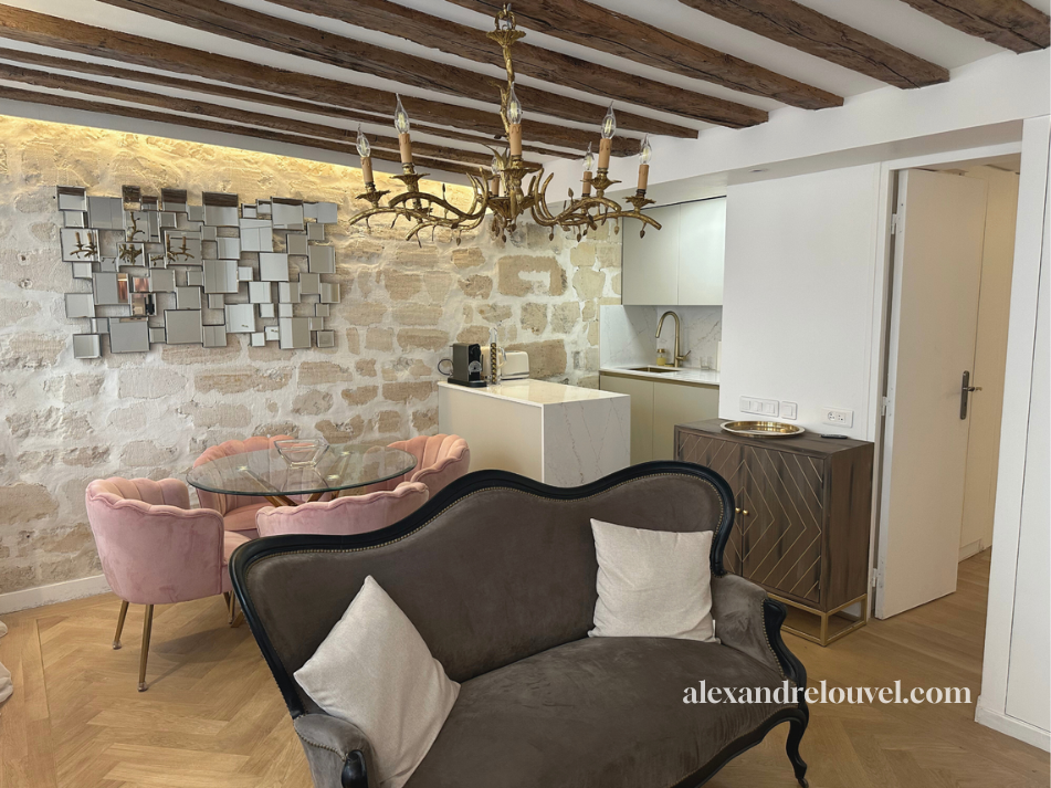 Salon avec canapé en velours gris, deux coussins blancs, table à manger en verre avec chaises pink, mur en pierre, miroir de décoration, plafond en bois, chandelier en métal, petite cuisine moderne en arrière-plan, sol en parquet