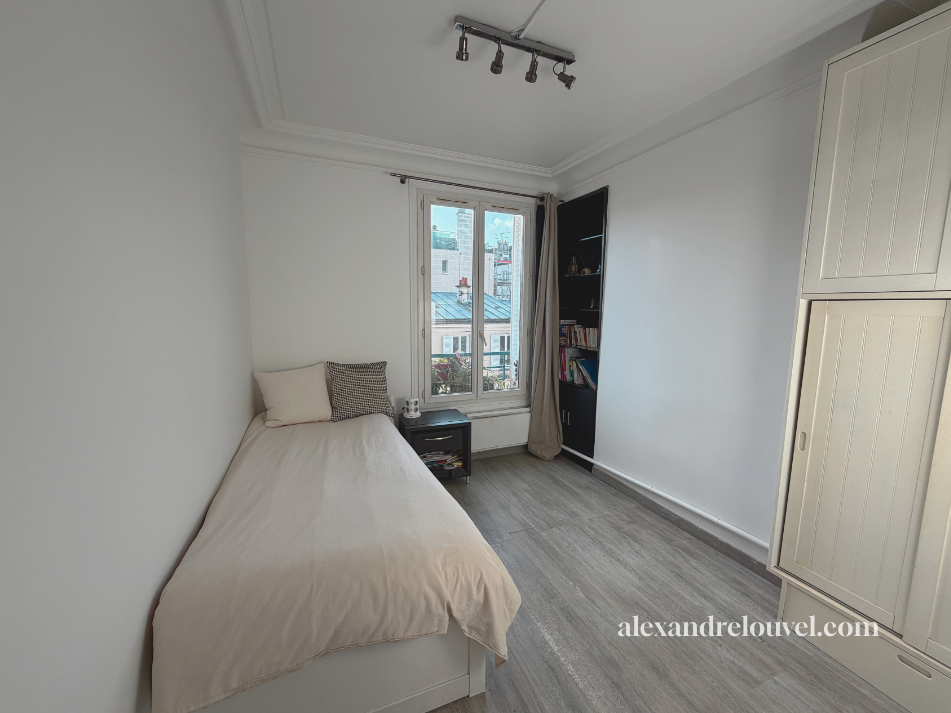 Chambre simple avec un lit contre le mur, une petite table de nuit, une fenêtre, une étagère noire avec des livres, un grand placard blanc, et un éclairage au plafond.