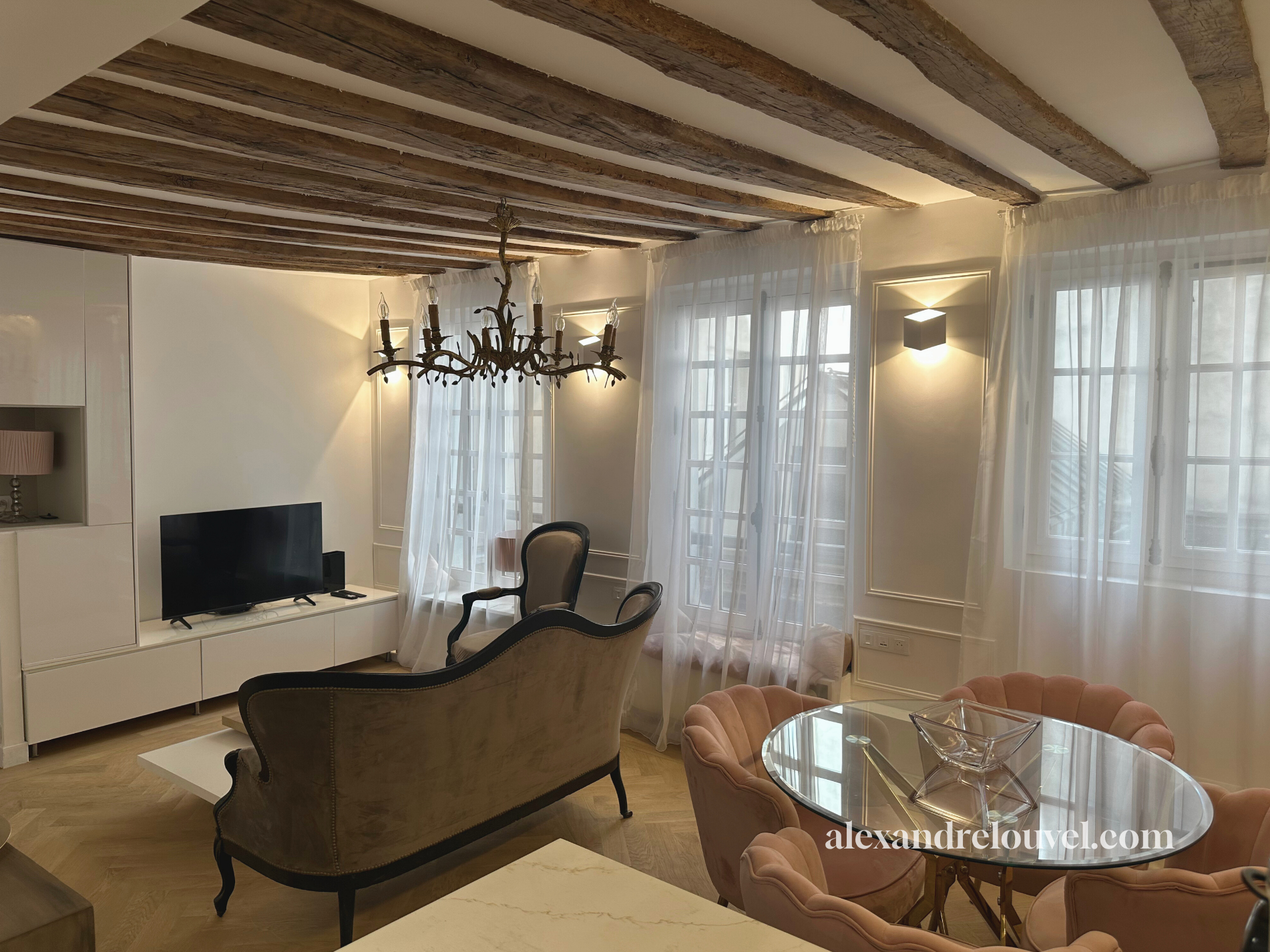 Salon avec meubles anciens, télévision, chandelier en métal noir, canapés en velours avec bois noir, table en verre avec sièges en velours rose, murs blancs, fenêtres avec rideaux légers, éclairage contemporain