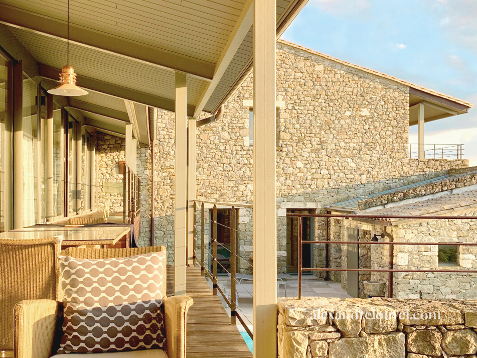 Balcon d'une maison en pierre avec mobilier en osier et vue sur un bâtiment en pierre similaire en arrière-plan.