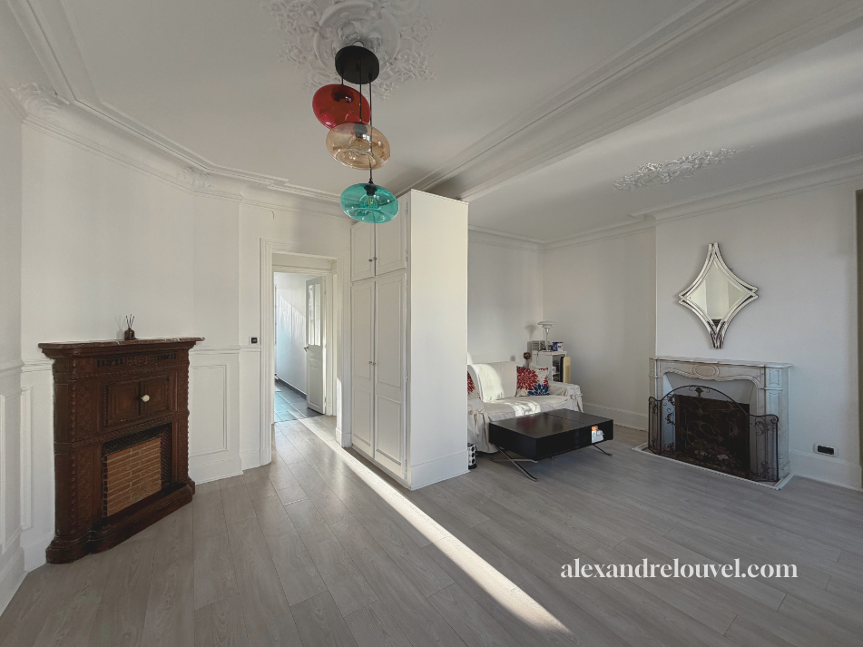 Salon avec cheminée en bois, canapé blanc avec coussins rouges, miroir ornemental au-dessus de la cheminée, plafond avec moulures, lampe suspendue colorée.