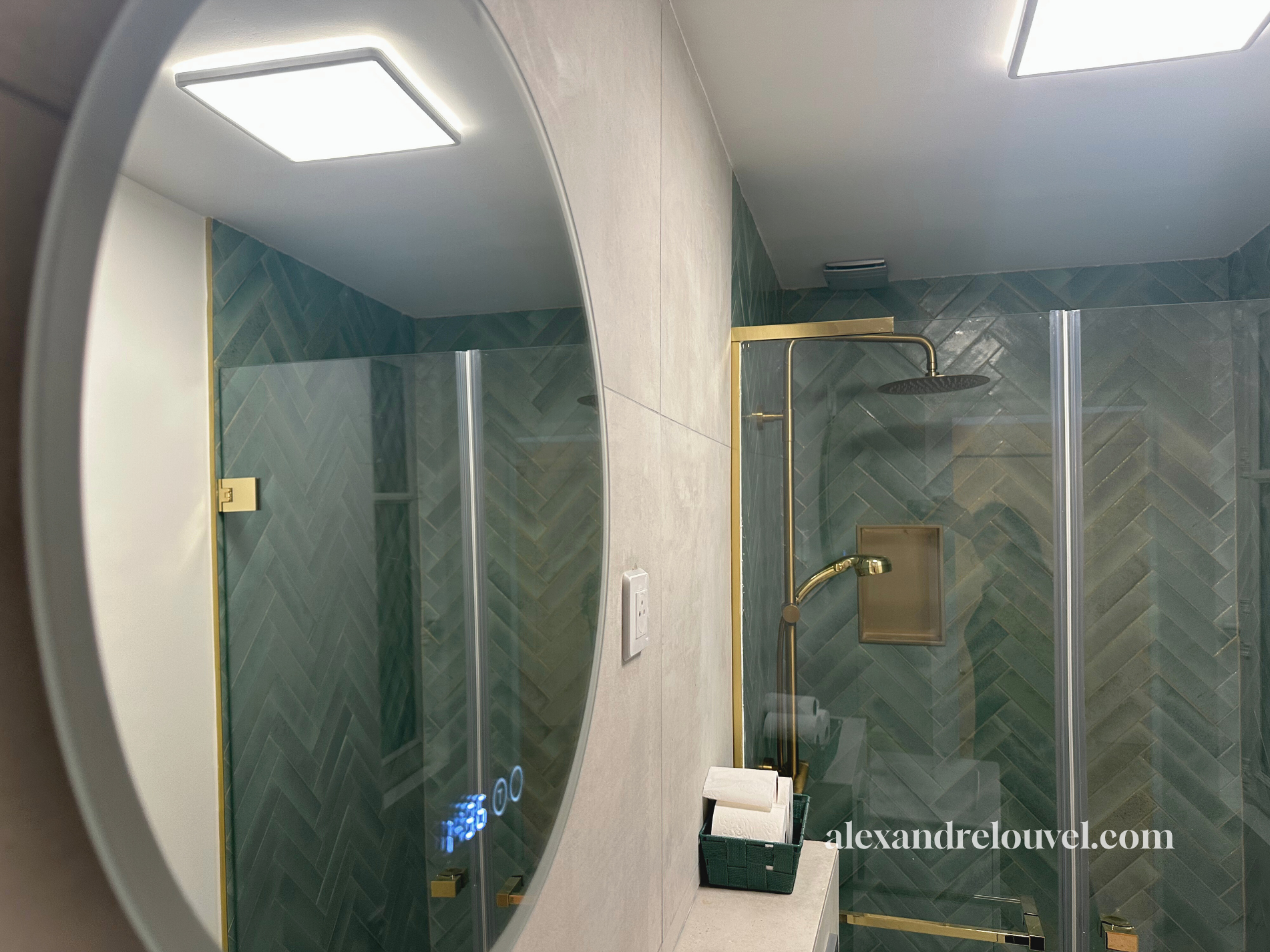 Salle de bain avec douche à l'italienne, carrelage gris en chevron, miroir, éclairage encastré, porte de douche en verre avec cadre doré, accessoires de toilette.