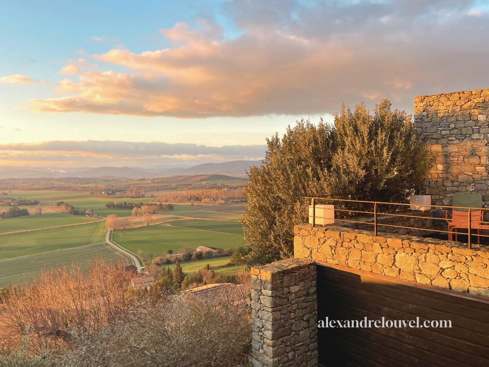 Vue d'un paysage champêtre au coucher du soleil avec des champs verts et des collines lointaines, vue depuis une terrasse en pierre avec une grande haie et un ciel orageux coloré.