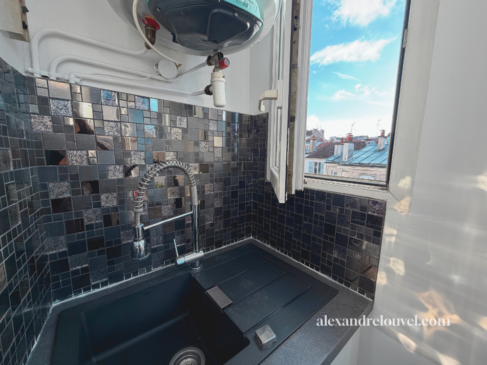 Petite cuisine ou coin cuisine avec un évier noir, un robinet extensible, un panneau de backsplash en carrelage noir brillant, une fenêtre ouverte donnant sur un ciel bleu avec quelques nuages et des toits d'immeubles. Logo de alexandra-louvel.com en