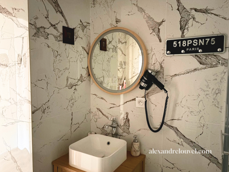 Salle de bain avec mur en marbre, miroir rond en bois, lavabo sur meuble en bois, sèche-cheveux fixé au mur, prise électrique, panneau Paris avec le code 518PSN75.