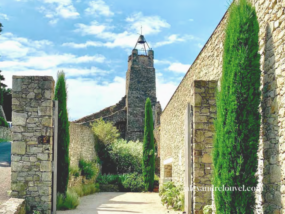 Une allée bordée de murs en pierre avec des arbres verts, menant à une vieille tour en pierre avec un ciel bleu en arrière-plan.