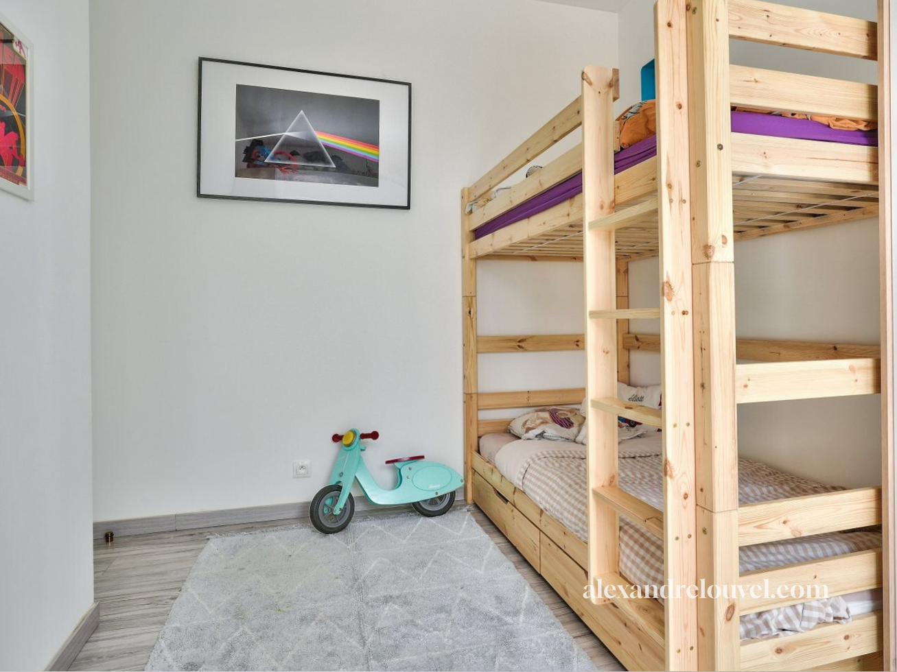 Une chambre d'enfant avec un lit superposé en bois, un tapis gris, un scooter en plastique vert et un tableau de Pink Floyd "The Dark Side of the Moon" accroché au mur.