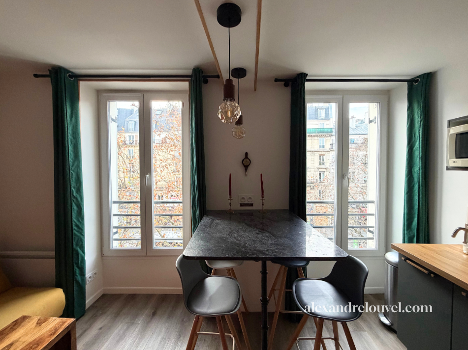 Intérieur d'une cuisine avec deux fenêtres, table en pierre, chaises, pendant, candélabres, horloge murale, et éléments de décoration.