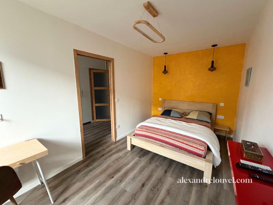 Chambre avec lit double, tête de lit en bois, deux lampes suspendues noires, un mur jaune derrière le lit, sol en bois, table de chevet en bois, meuble rouge avec télécommande et boîte, porte ouverte menant à un autre espace, plafond blanc avec éclai