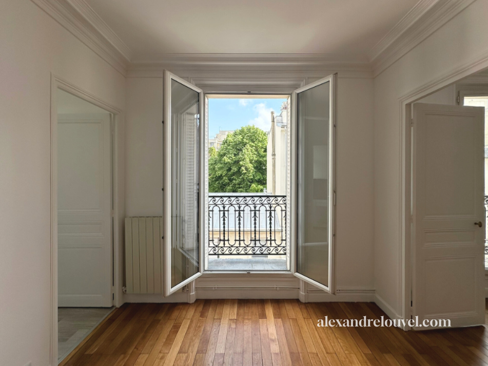 Une chambre lumineuse avec des fenêtres ouvertes donnant sur un balcon en fer forgé et une vue sur des arbres et des bâtiments.