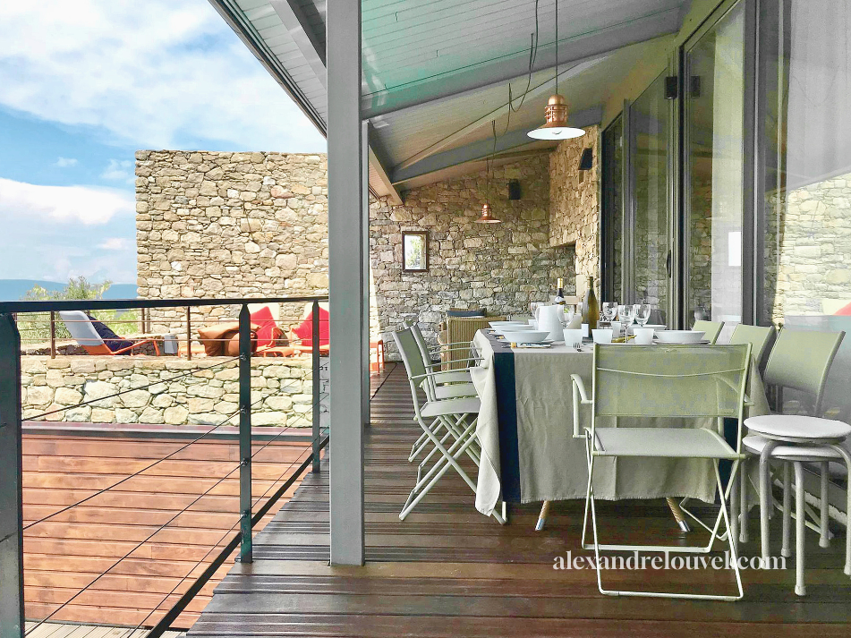 Une terrasse avec une table à manger dressée à l'intérieur entourée de chaises, vue sur un ciel bleu et un mur en pierre, avec des fauteuils sur une terrasse extérieure en pierre.
