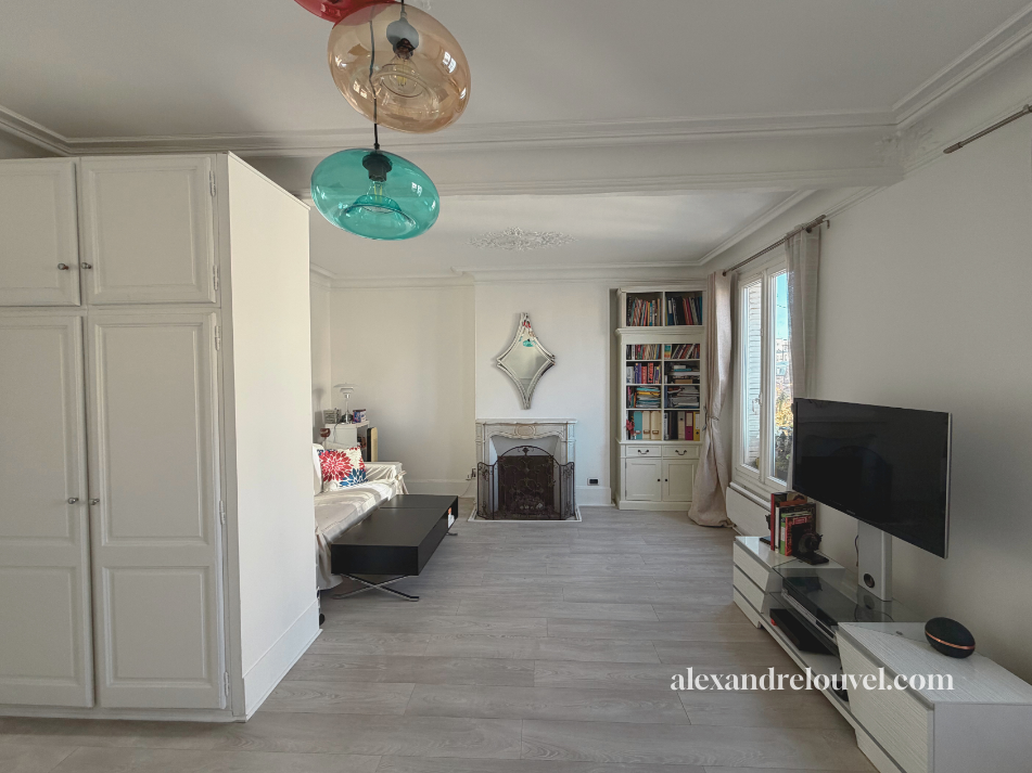 Salon avec canapé blanc, cheminée, étagère avec des livres, télévision, et luminaires colorés suspendus au plafond.