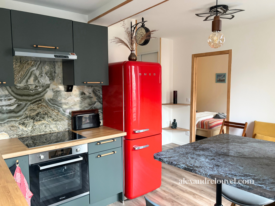 Cuisine avec un réfrigérateur rouge SMEG, un four, une plaque de cuisson, des armoires grises, un plan de travail en bois, et un mur en marbre. Il y a aussi une table de salle à manger en pierre et un salon avec un canapé jaune en arrière-plan.