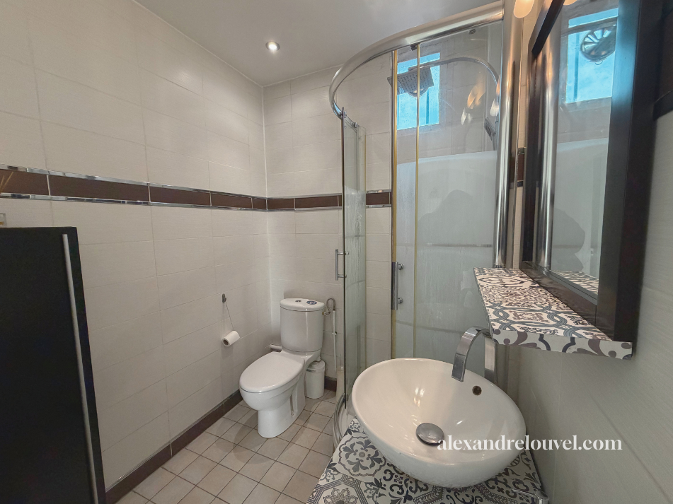 Petite salle de bain avec une toilette, un lavabo, une douche en coin, et un miroir avec étagère decorative, sol en carrelage beige, murs carrelés blancs avec une ligne marron.