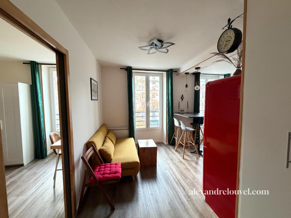 Salon lumineux avec canapé jaune, table en bois, fauteuil rouge, fenêtre avec rideaux verts, cuisine en arrière-plan avec île et chaises, horloge murale noire, décor avec plantes et lampes suspendues.