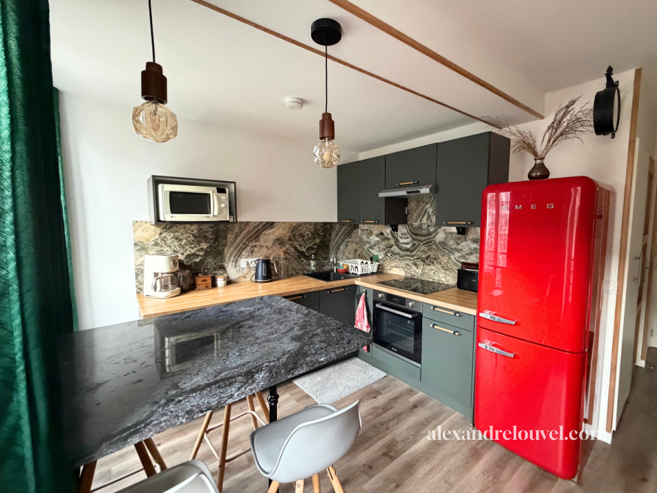 Cuisine moderne avec meubles gris, plan de travail en bois, réfrigérateur rouge vintage, électroménagers noirs et en acier, éclairages suspendus, mur en marbre, vaisselle, tasse, micro-ondes, plante en vase, chaise blanche.