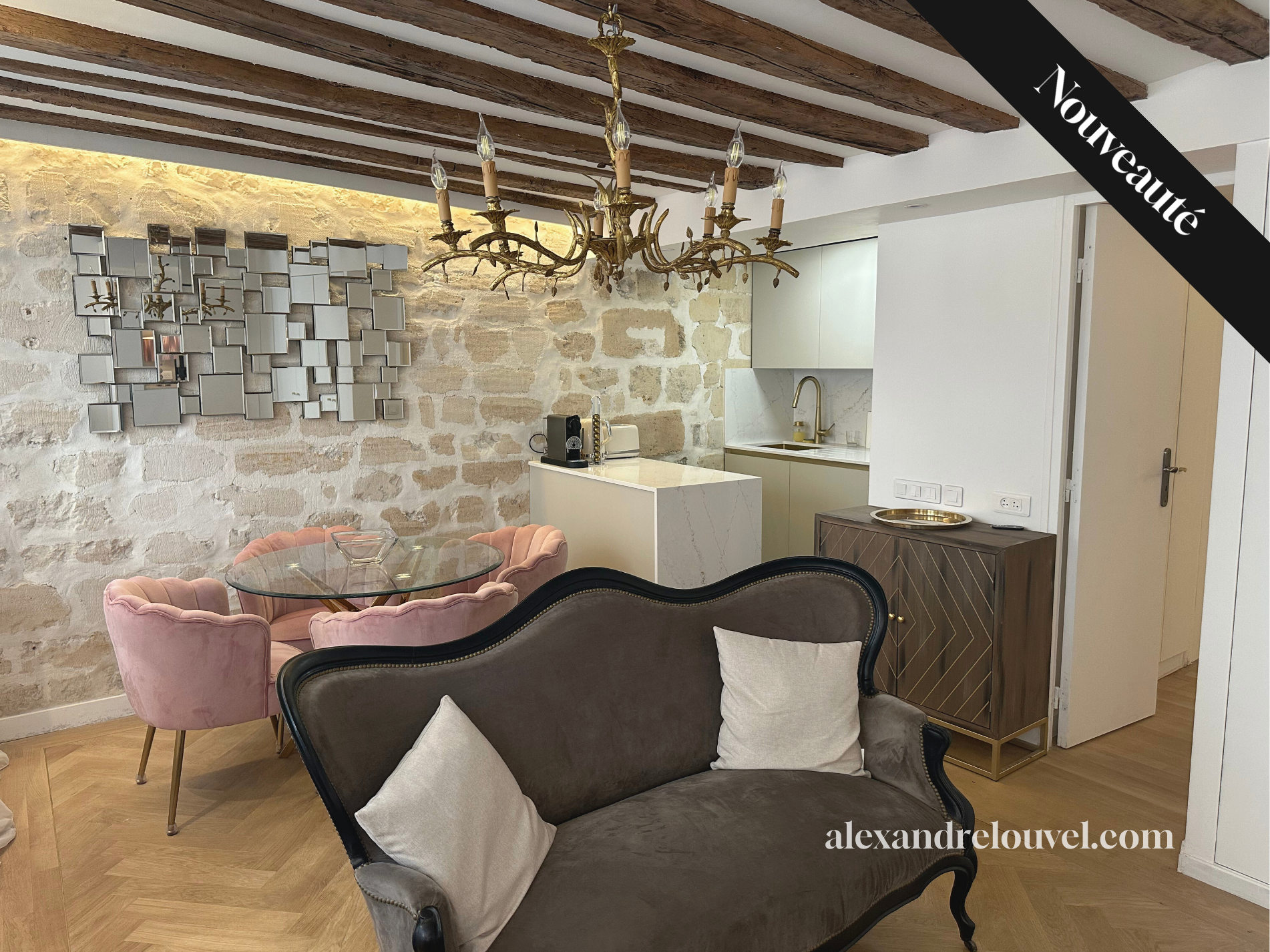 Salon avec canapé ancien, table en verre, chaises roses en velours, plafond en bois, mur en pierre, miroir en mosaïque, chandelier en métal doré, petite cuisine blanche avec évier doré, meuble en bois avec plateau doré, porte blanche.