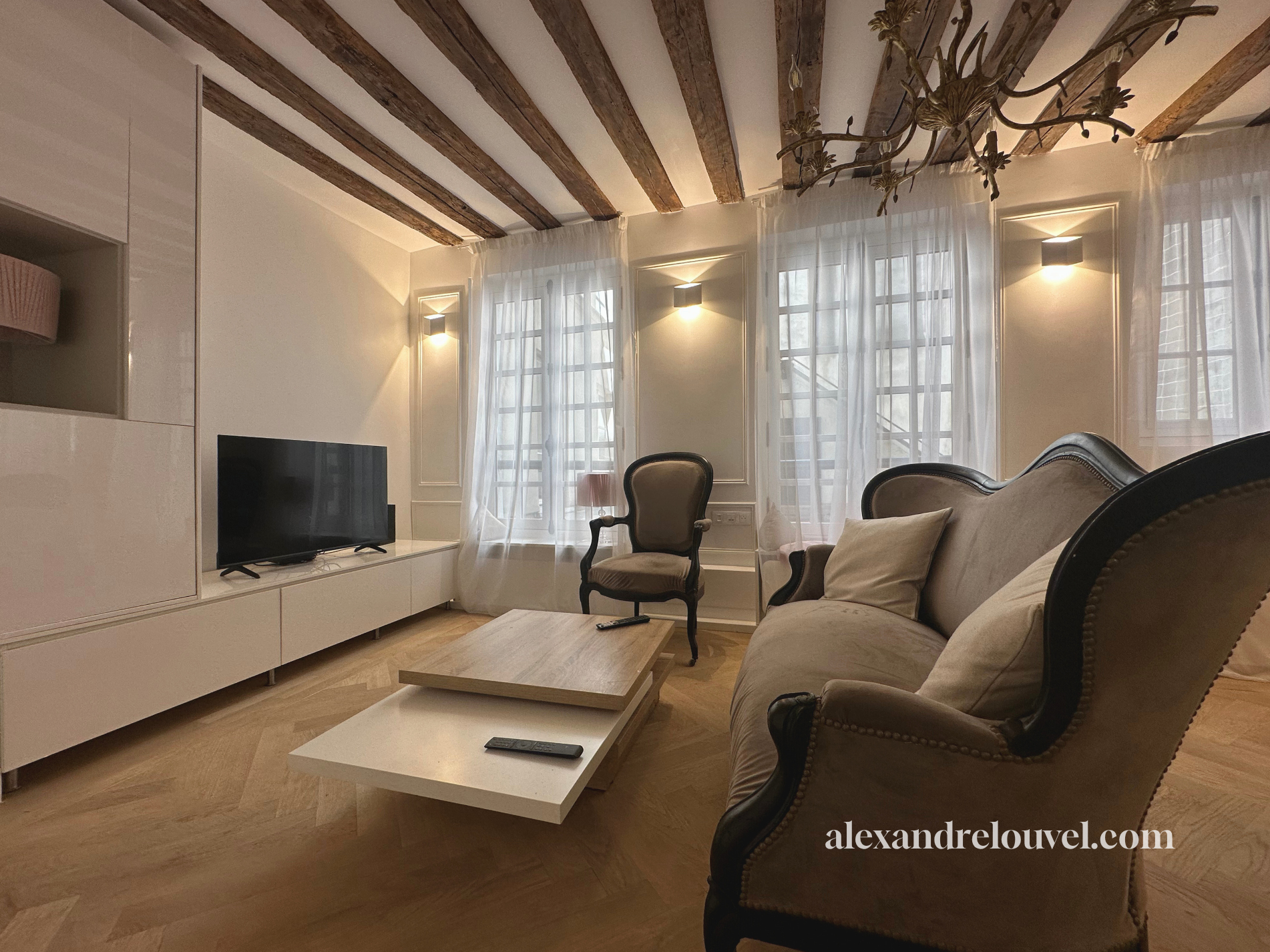 Salon moderne avec canapé beige, fauteuil, télévision sur meuble blanc, grande fenêtre avec rideaux transparents, plafonnier en chandelier, parquet en bois, éclairage mural