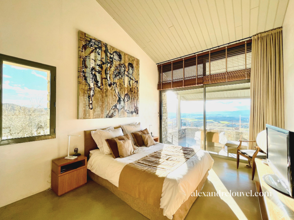 Chambre avec un lit king-size, tableau abstrait au-dessus, fenêtre avec vue sur la nature, porte-fenêtre donnant sur un balcon avec vues panoramiques, meubles en bois et rideaux beiges.
