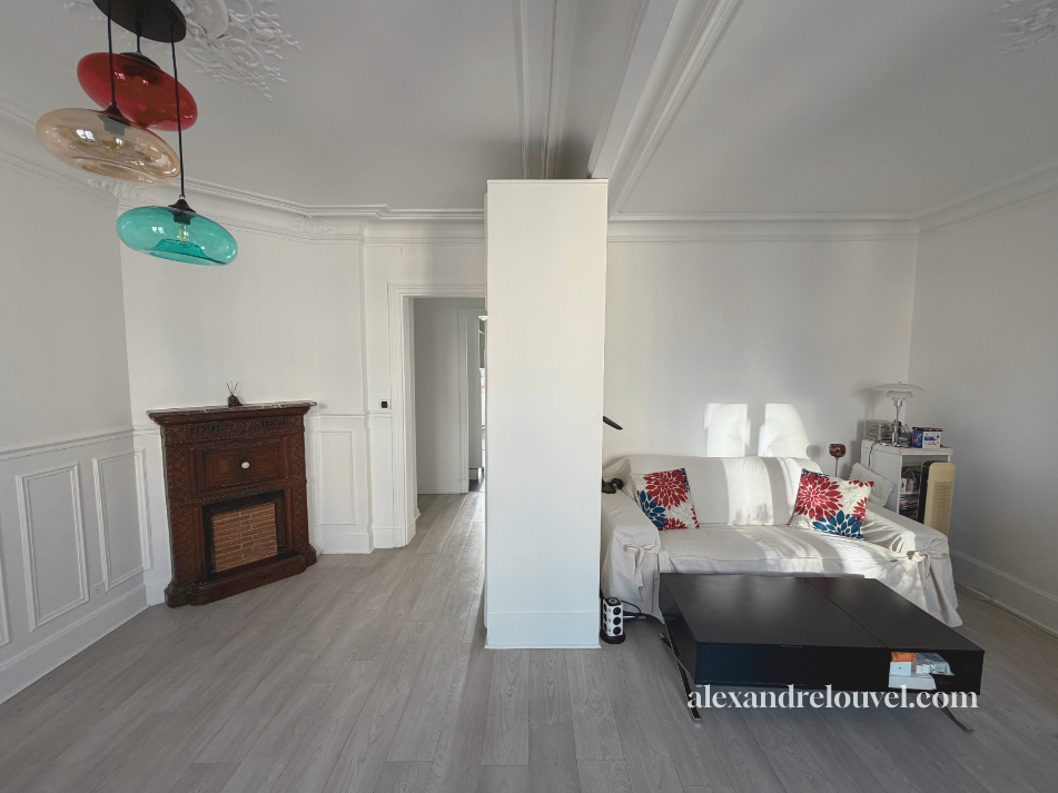 Salon avec un canapé blanc, une table basse noire, une cheminée ancienne en bois, des coussins rouges à motifs floraux, et un coin lumineux avec une lampe et une étagère. Trois pendentifs colorés sont suspendus au plafond.