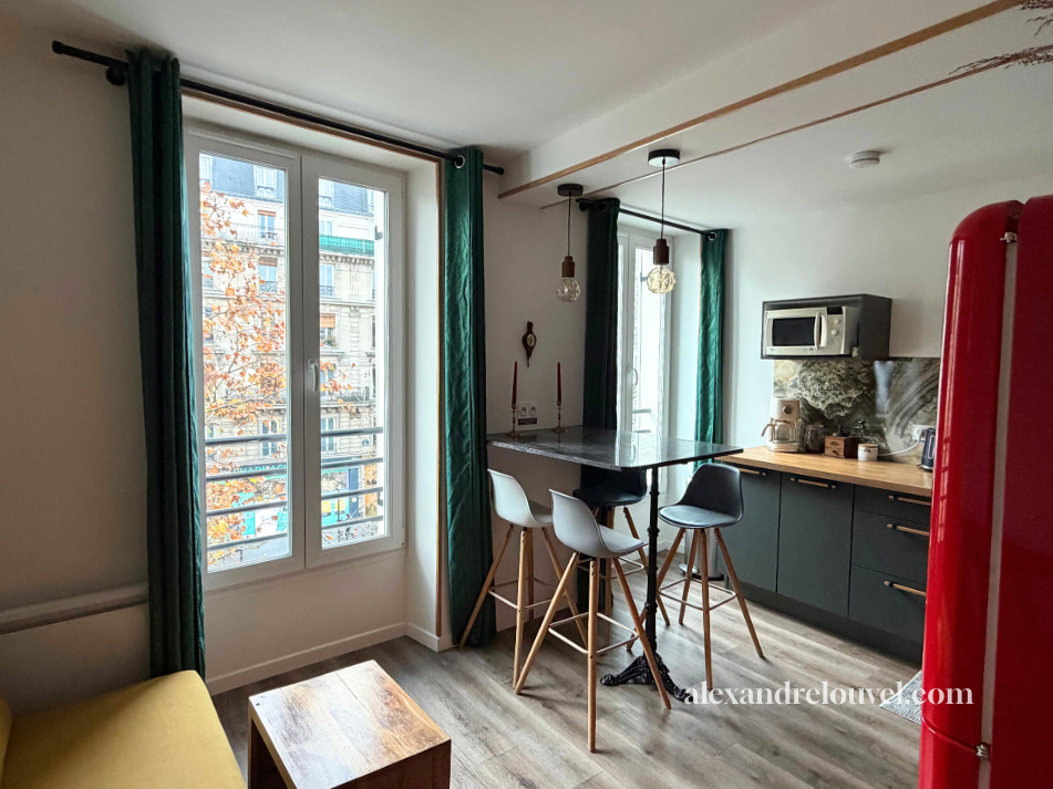 Cuisine moderne avec fenêtre, table centrale, chaises de bar, micro-ondes, réfrigérateur rouge, décoration minimaliste.