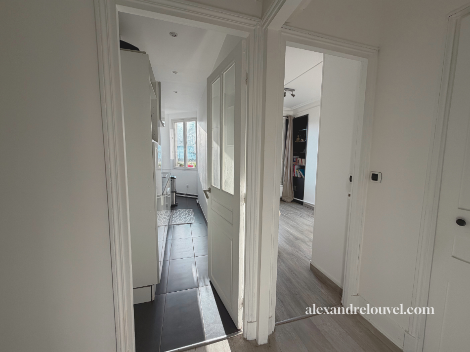 Vue d'une entrée dans une cuisine et un salon d'une maison avec murs blancs, sol en carrelage dans la cuisine, et sol en bois dans le salon. La cuisine dispose d'une fenêtre qui laisse entrer la lumière naturelle.