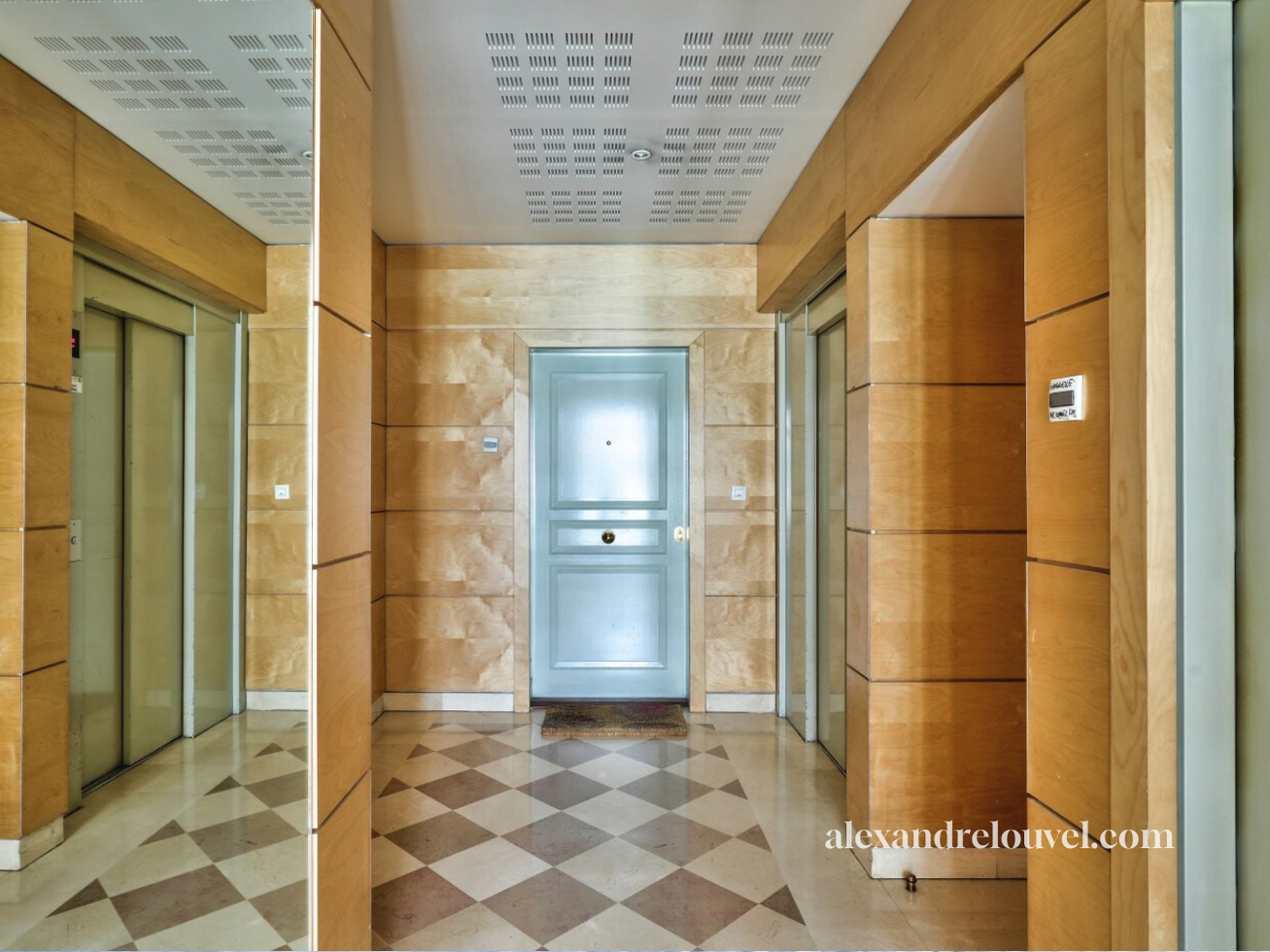 Hall d'entrée avec trois ascenseurs, mur en bois clair, sol en carreaux beige et marron, porte en métal au fond, tapis devant la porte, site alexandrelouvel.com indiqué en bas à droite.