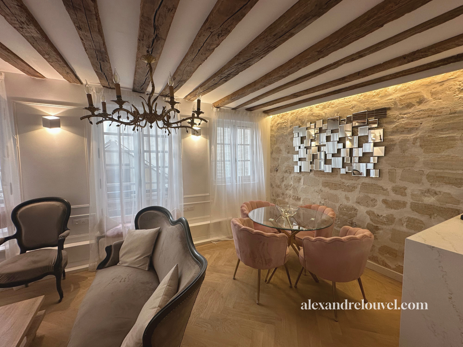 Salon avec chandelle en bois, canapé, fauteuils, table en verre, murs en pierre, miroir moderne, fenêtres avec rideaux blancs, éclairage tamisé