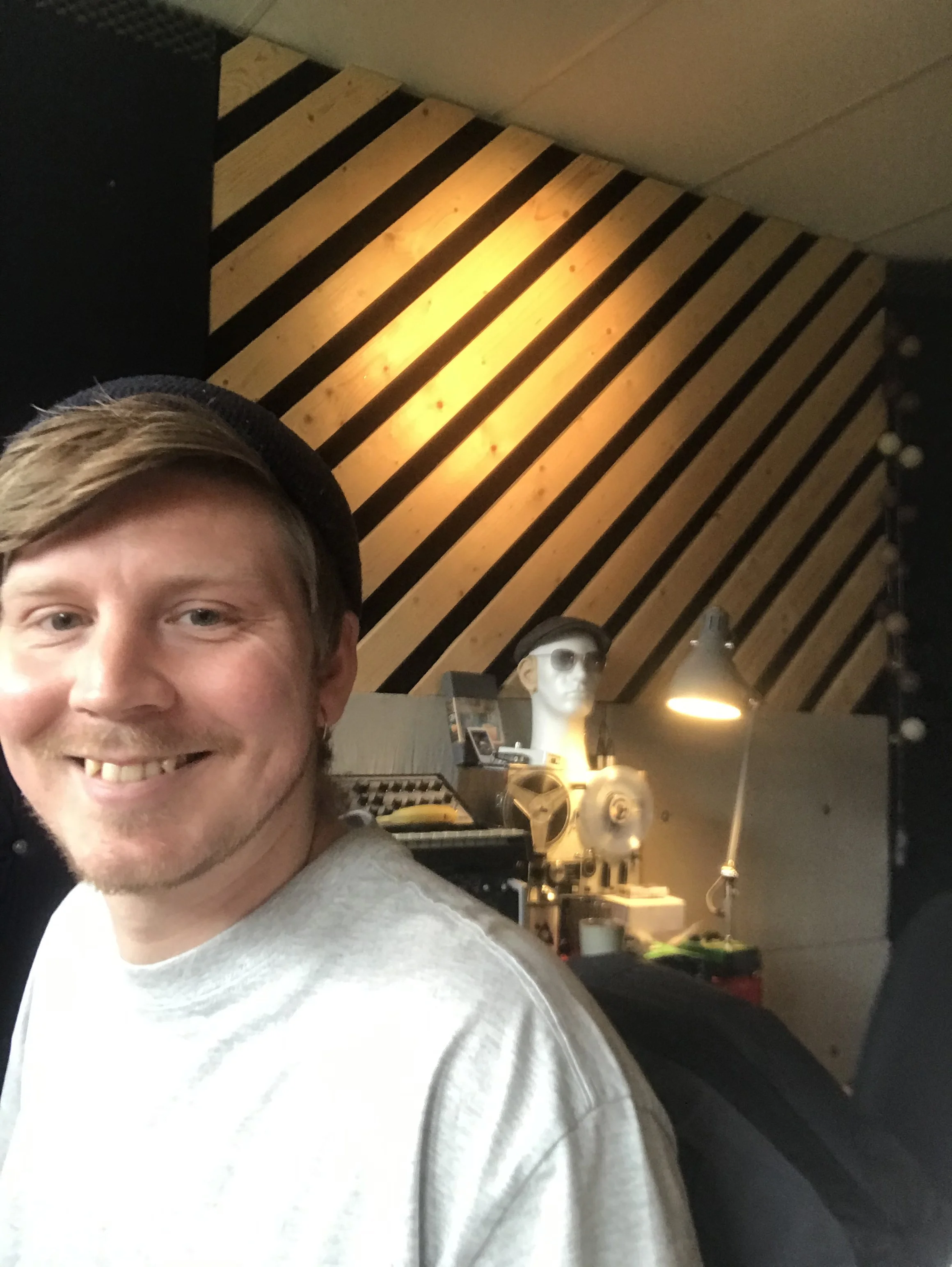 Ung mand med lyst hår, smilende, i et rum med væg af træpaneler og studieudstyr | Musikkomposition | Lyddesign | Filmproduktion Aarhus | GESTALT