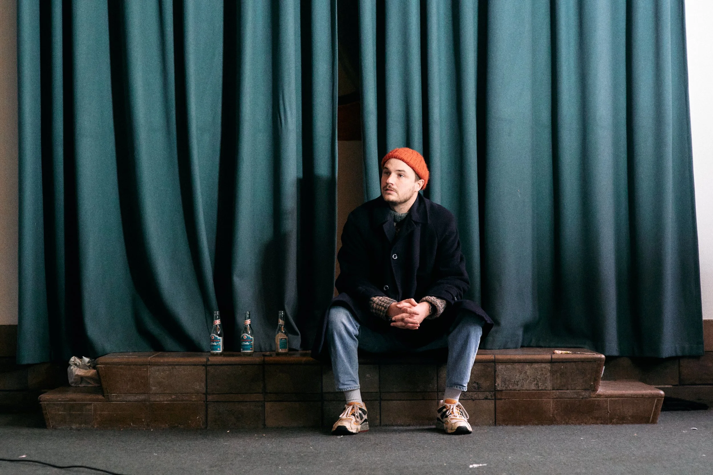 En mand sidder på trappetrin foran en grøn gardin, iført en orange hue, sort jakke, blå jeans og sneakers, med tre brugte glasflasker ved siden af ham | Dokumentarfilm | Retrograd | Esben Persson & Anders Mielcke | Filmproduktion Aarhus | GESTALT