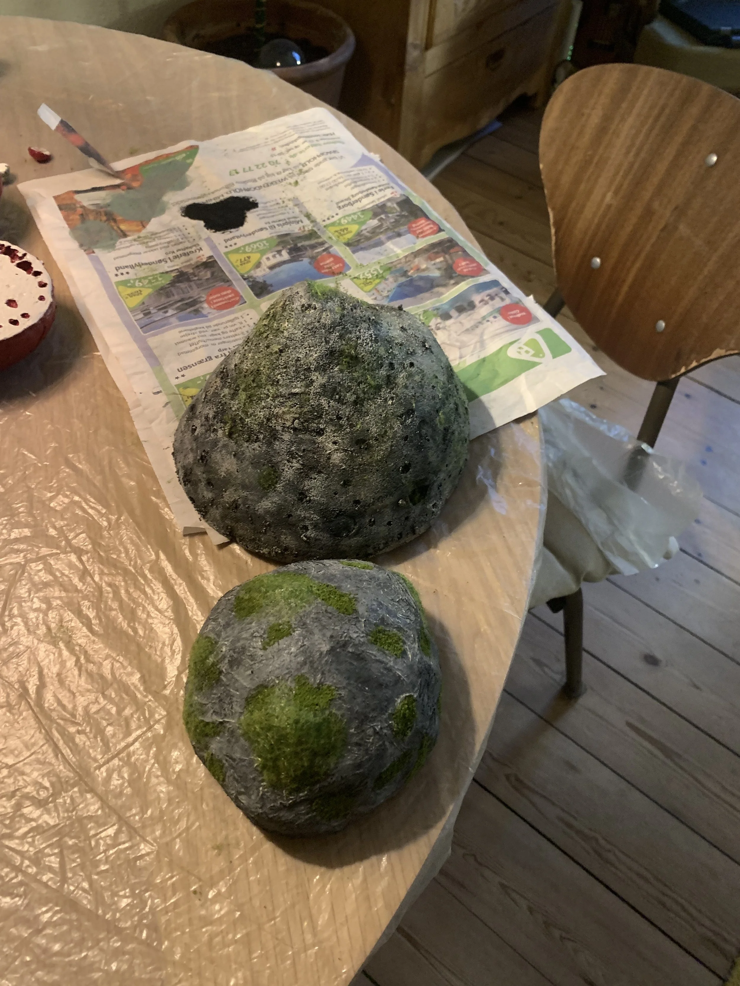 To store kunstige sten, en større med grøn mos og en mindre med mørke pletter, på et bord dækket med papir og aviser | Dokumentarfilm | Retrograd | Esben Persson & Anders Mielcke | Filmproduktion Aarhus | GESTALT