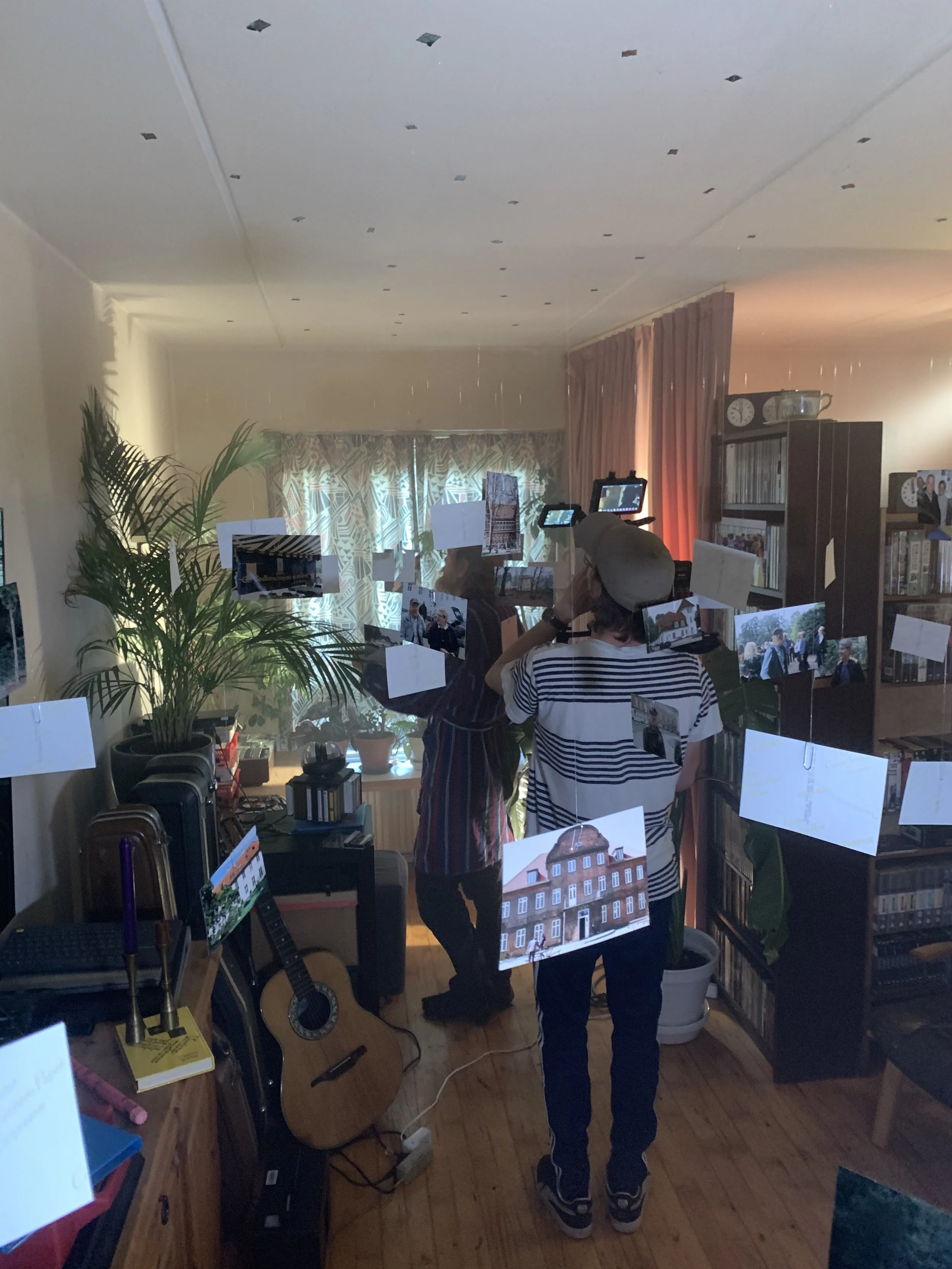 Indendørs scene med to personer og kamera, omgivet af fotos hængt op i et rum med bogreoler og planter | Dokumentarfilm | Retrograd | Esben Persson & Anders Mielcke | Filmproduktion Aarhus | GESTALT