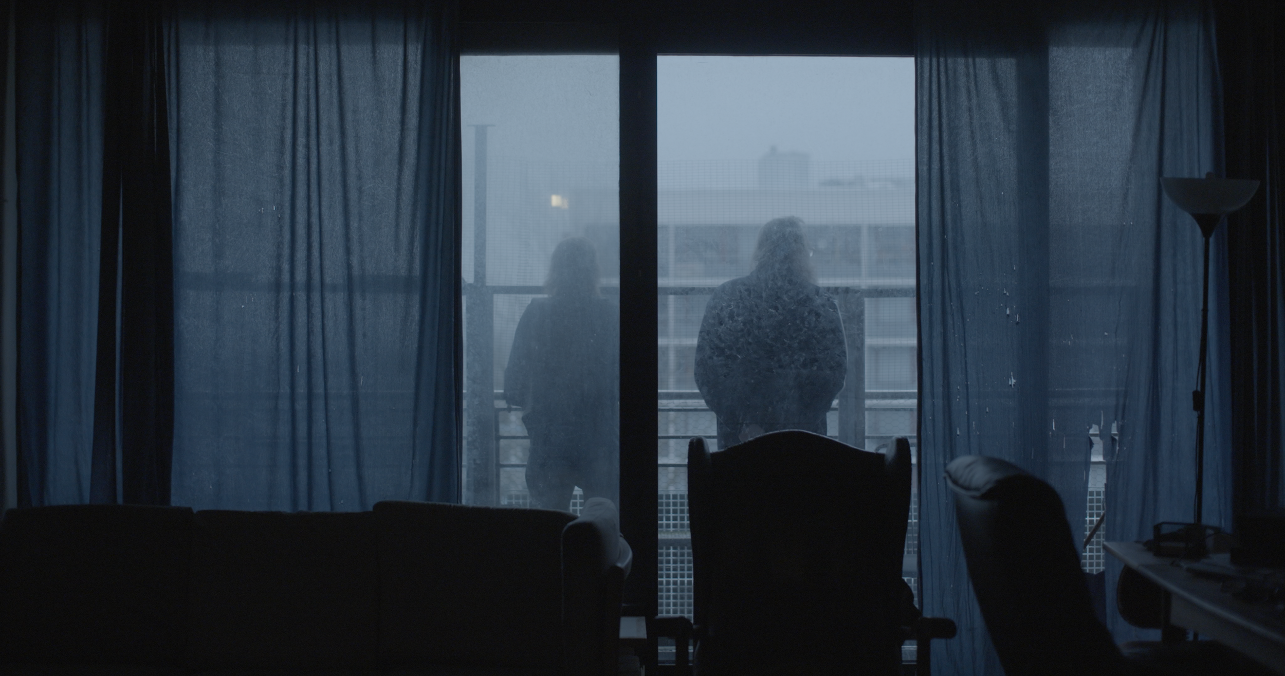 To personer står på en balkon set fra indersiden af en lejlighed, gennem en glasdør med blå gardiner, hvor det er dystert og regnfuldt udenfor | Dokumentarfilm | Retrograd | Esben Persson & Anders Mielcke | Filmproduktion Aarhus | GESTALT