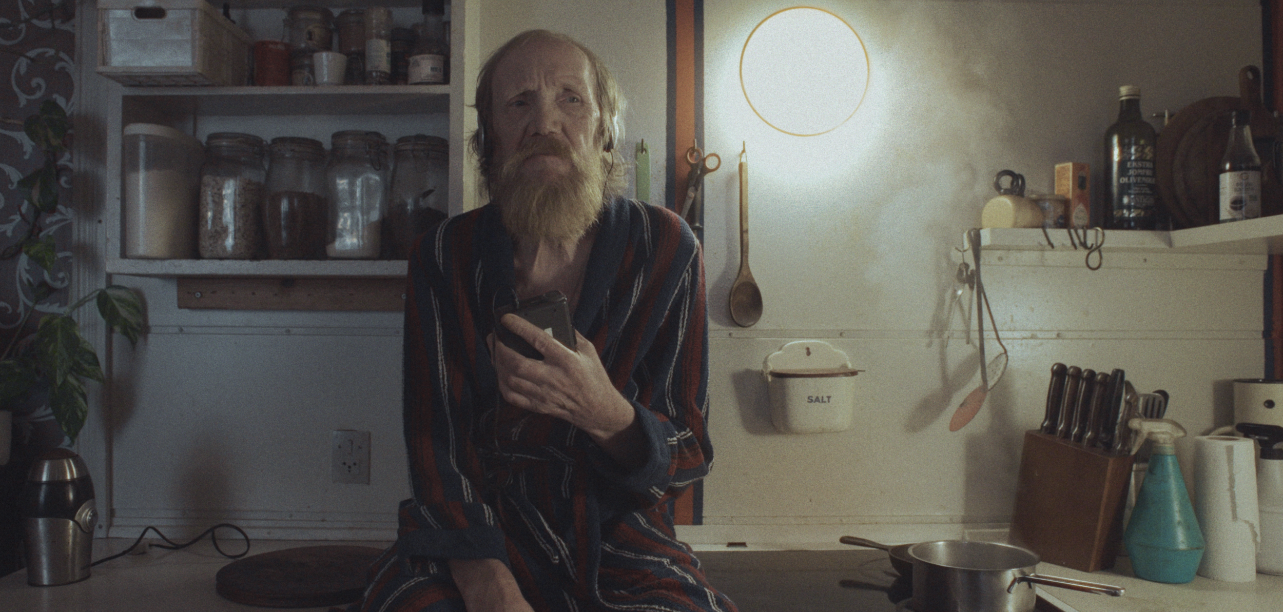 En ældre mand med lang hår og skæg sidder i et køkken iført en stribet badekåbe | Dokumentarfilm | Retrograd | Esben Persson & Anders Mielcke | Filmproduktion Aarhus | GESTALT