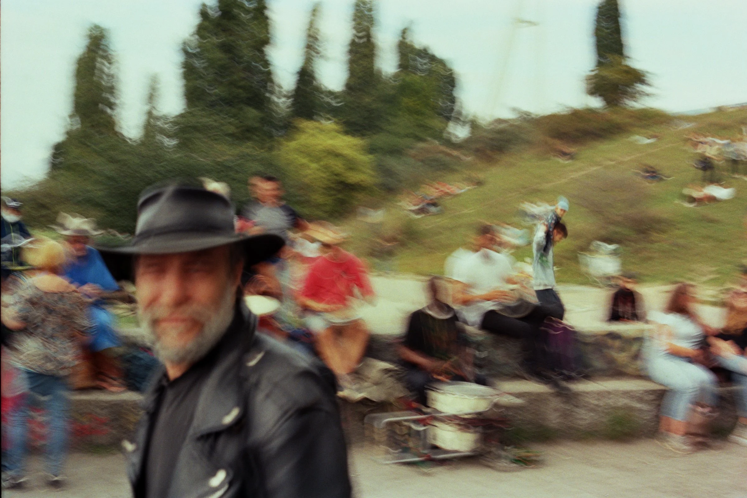 En mand med hat i forgrunden, omkranset af personer, der sidder på en sten og forbipasserende, i en udendørs park eller strand | Location scouting Berlin | Kodak | Filmproduktion Aarhus | GESTALT