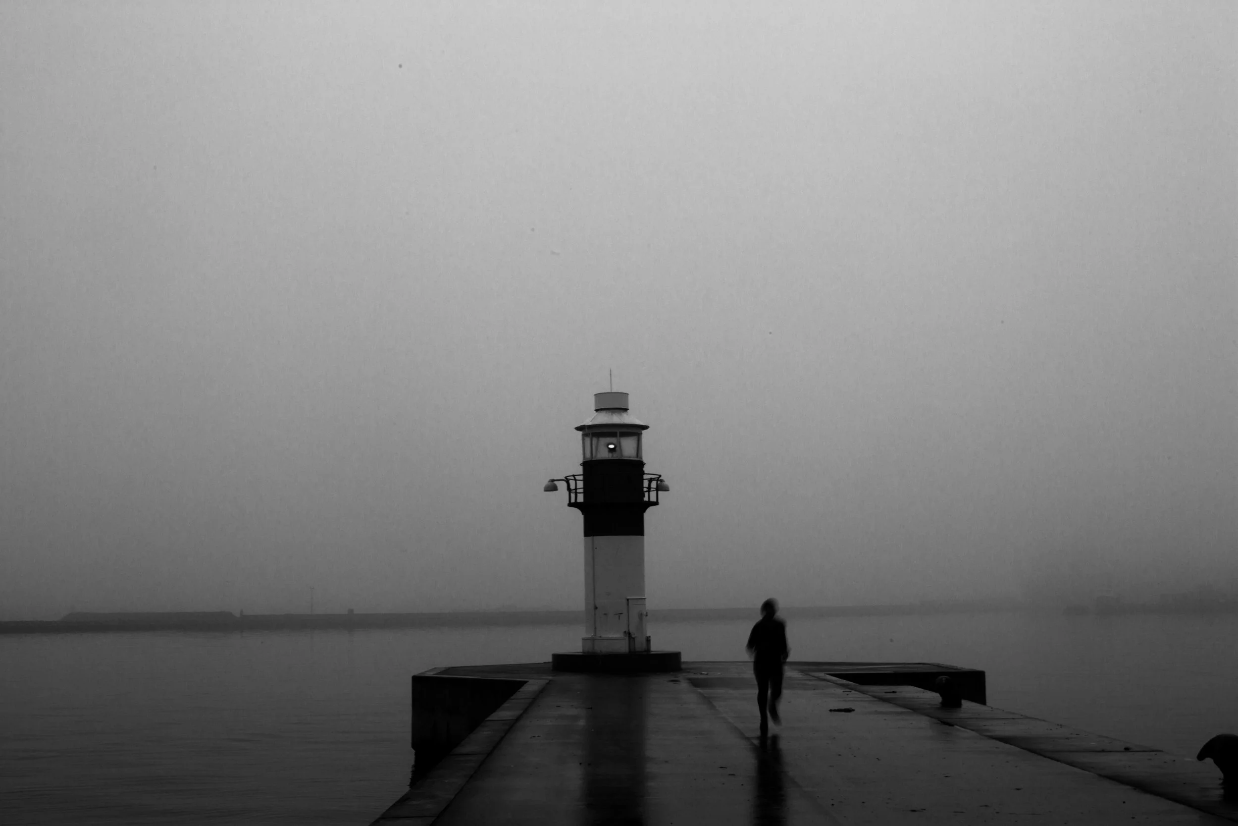 Sort-hvid billede af en person, der går på en mole mod en fyrtårn ved vandet, med en overskyet himmel | Location scouting | Kodak | Filmproduktion Aarhus | GESTALT