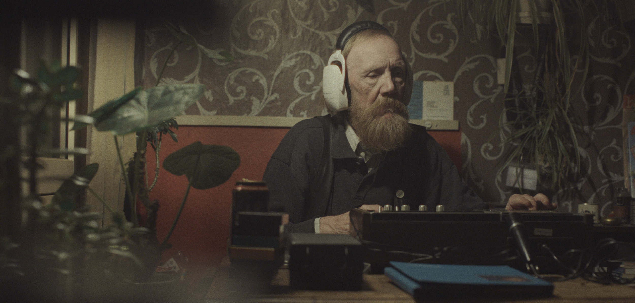 En mand med skæg, der sidder ved et skrivebord med høretelefoner, omgivet af planter | Dokumentarfilm | Retrograd | Esben Persson & Anders Mielcke | Filmproduktion Aarhus | GESTALT