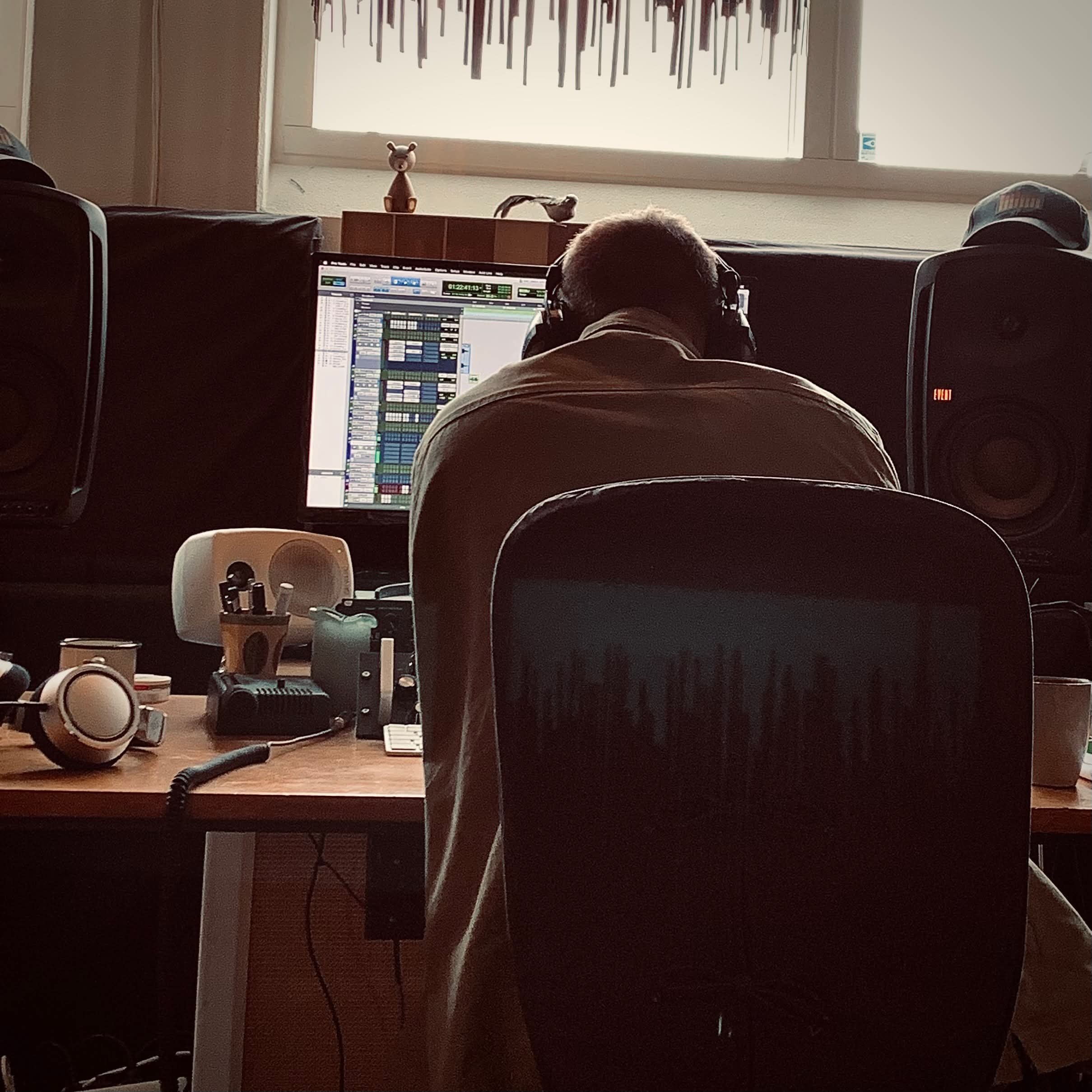 Person sidder foran en computerskærm med lyd- og musiksoftware, omgivet af professionel lydudstyr i et hjemmestudie, med en solbeskinnet vindue i baggrunden | Musikkomposition | Studieproduktion og lyddesign | Filmproduktion Aarhus | GESTALT