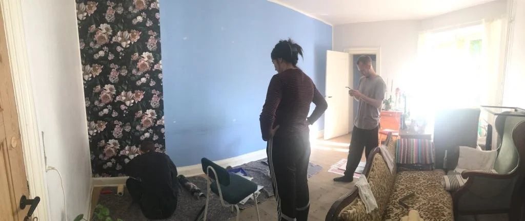 Tre personer i stue, to står og en sidder på gulvet, møbler og blomstertapet sættes på væggen | Kortfilm | In Medias Ræs | Esben Persson | Filmproduktion Aarhus | GESTALT