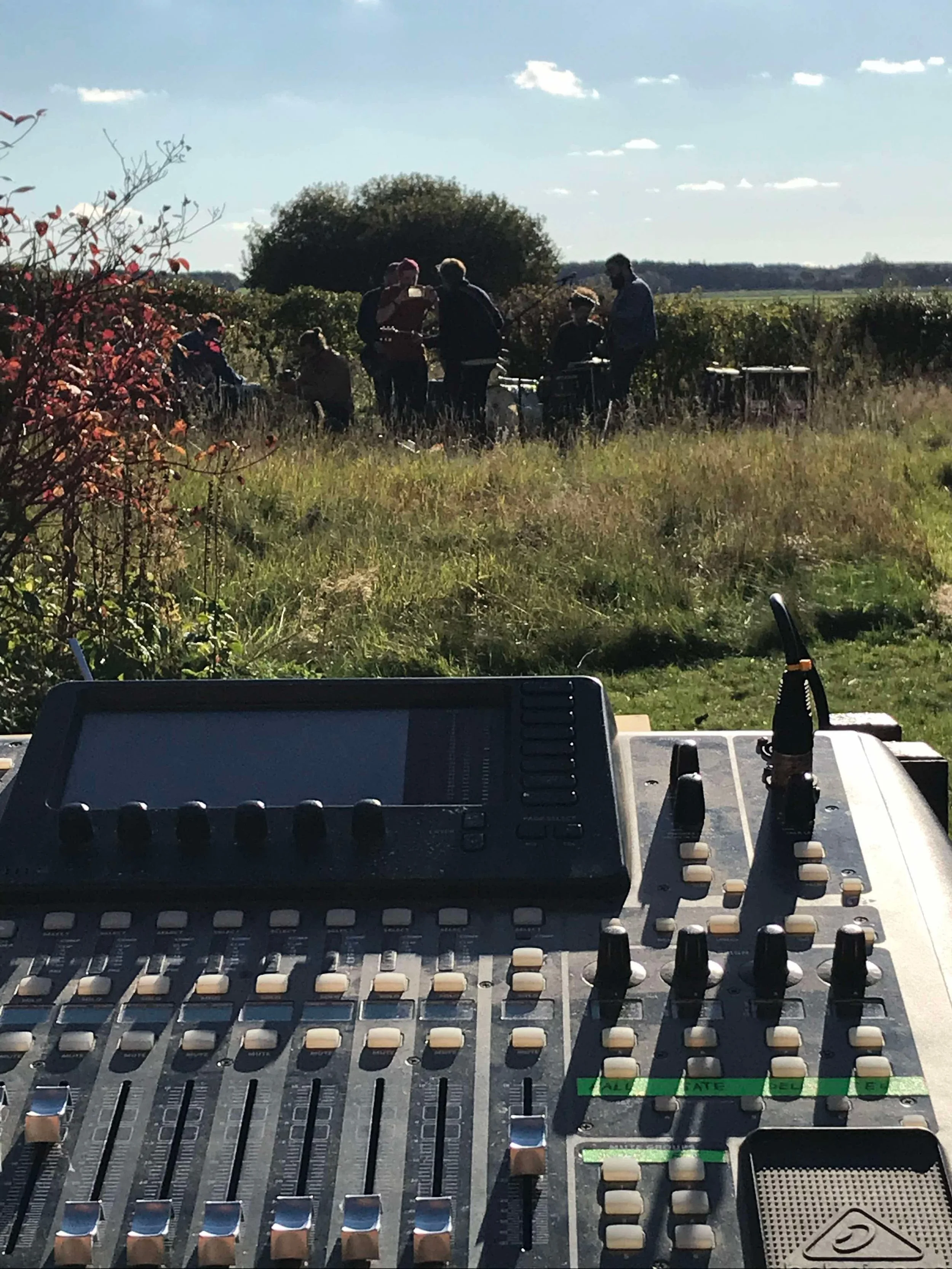 Udenfor i et åbent område er der en gruppe mennesker, der står og sidder i et solrigt landskab, set bagfra. I forgrunden ses et lydsetup | Studieproduktion | Musikkomposition | Filmproduktion Aarhus | GESTALT
