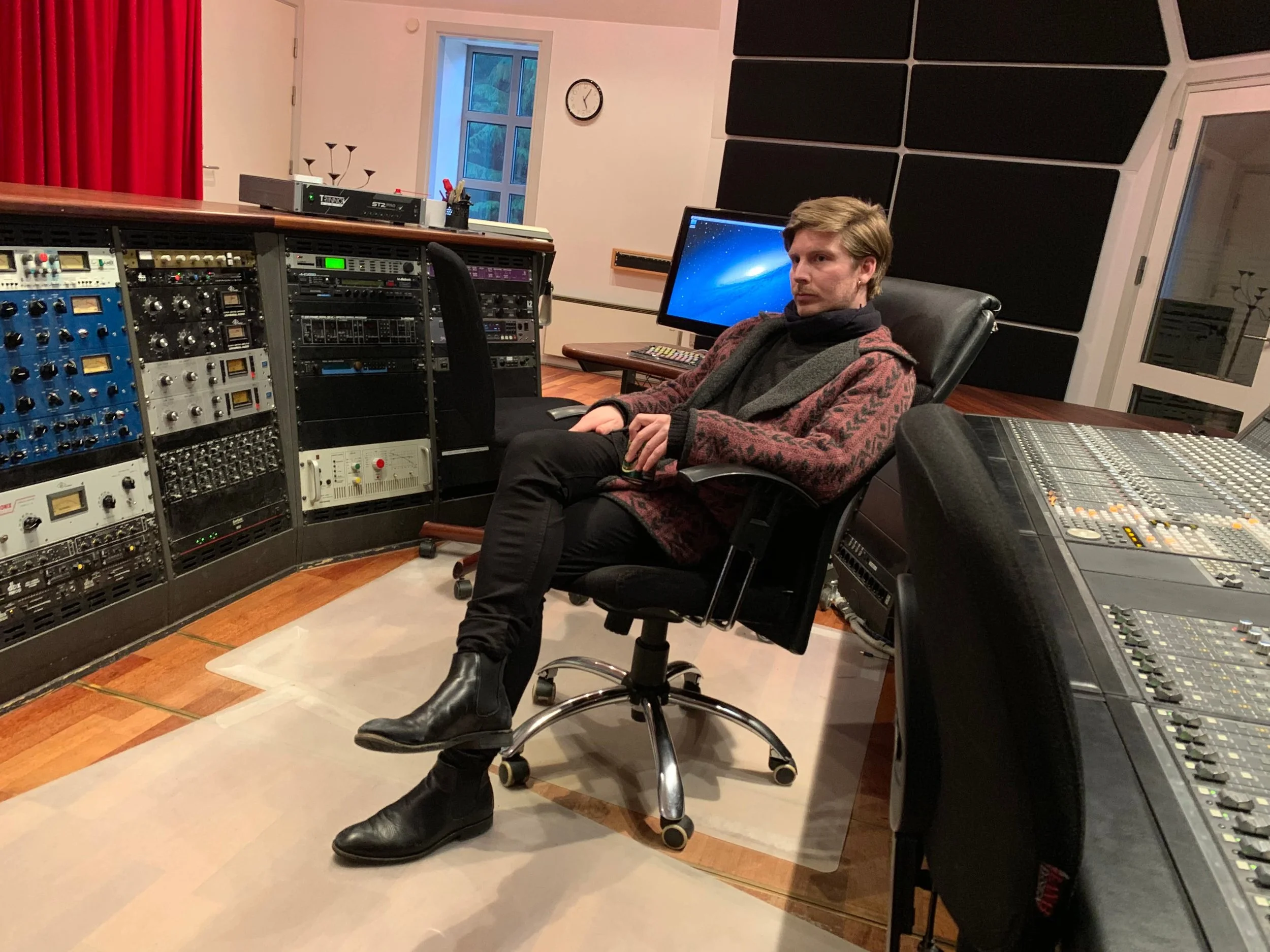 En ung mand sidder i en lydteknikerstol i et lydstudie med stor lydmixer på bordet til højre og forskellige lydracks foran ham. Bag ham er der en computerskærm | Musikkomposition | Studieproduktion og lyddesign | Filmproduktion Aarhus | GESTALT