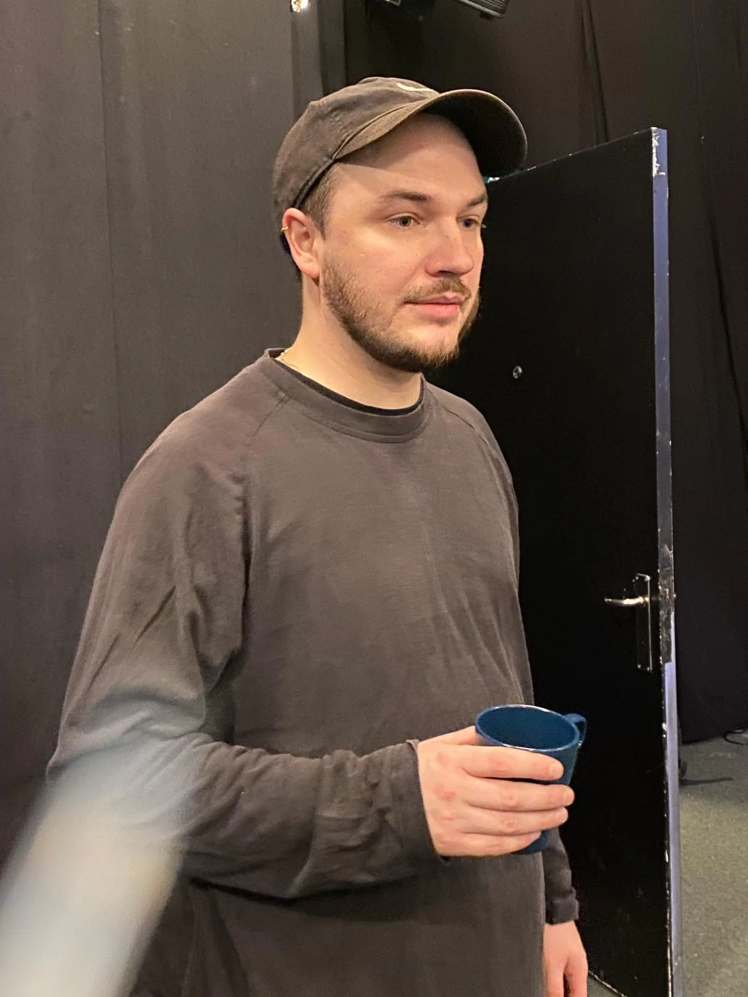 Ung mand med brunt hår og skæg i gråt sweatshirt og sort hat holder en blå kop, står foran en sort væg og dør | Dokumentarfilm | Retrograd | Esben Persson & Anders Mielcke | Filmproduktion Aarhus | GESTALT