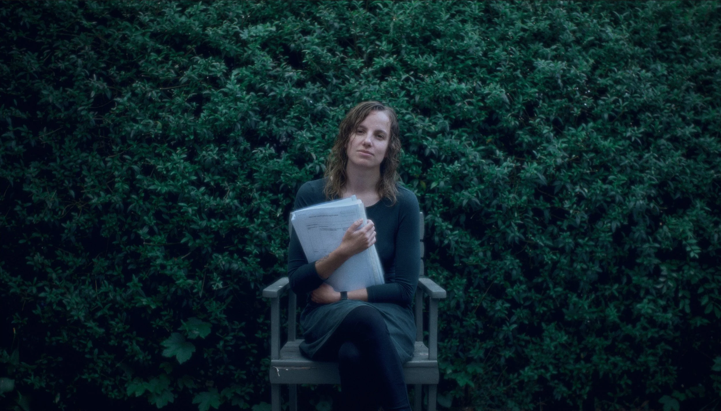 En kvinde sidder på en stol foran en grøn busk, holder en bunke papirer | Dokumentarfilm | Retten til egen historie | Anders Mielcke Grønbæk | Filmproduktion Aarhus | GESTALT