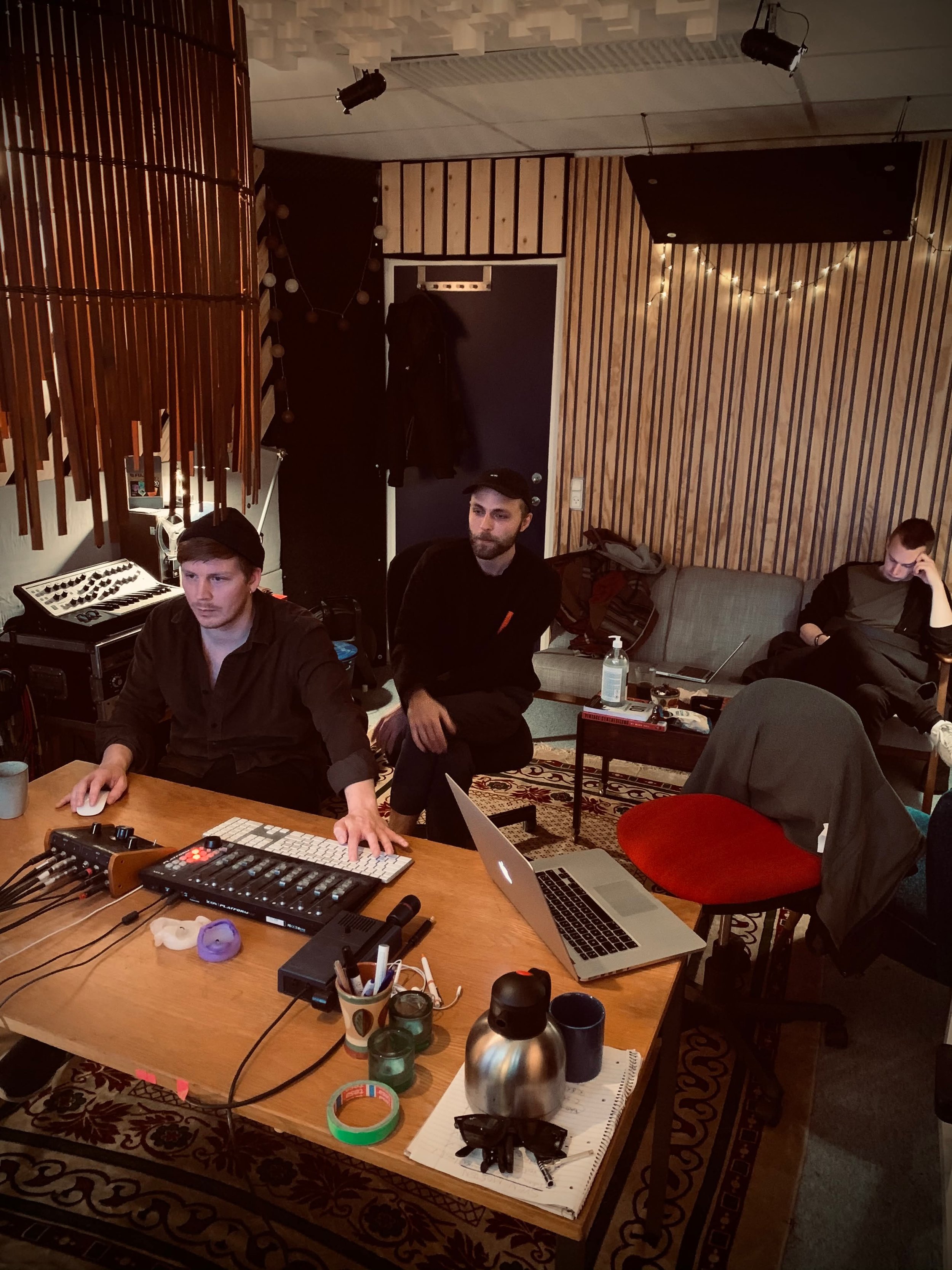 Tre mænd i et musikstudiolokale med lydudstyr og laptops, to sidder ved et bord og en sidder på en sofa bagved | Musikkomposition | Studieproduktion og lyddesign | Filmproduktion Aarhus | GESTALT